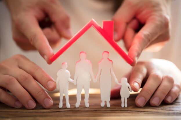 Two People Protecting Family Figures With Red Roof Over Wooden Desk
